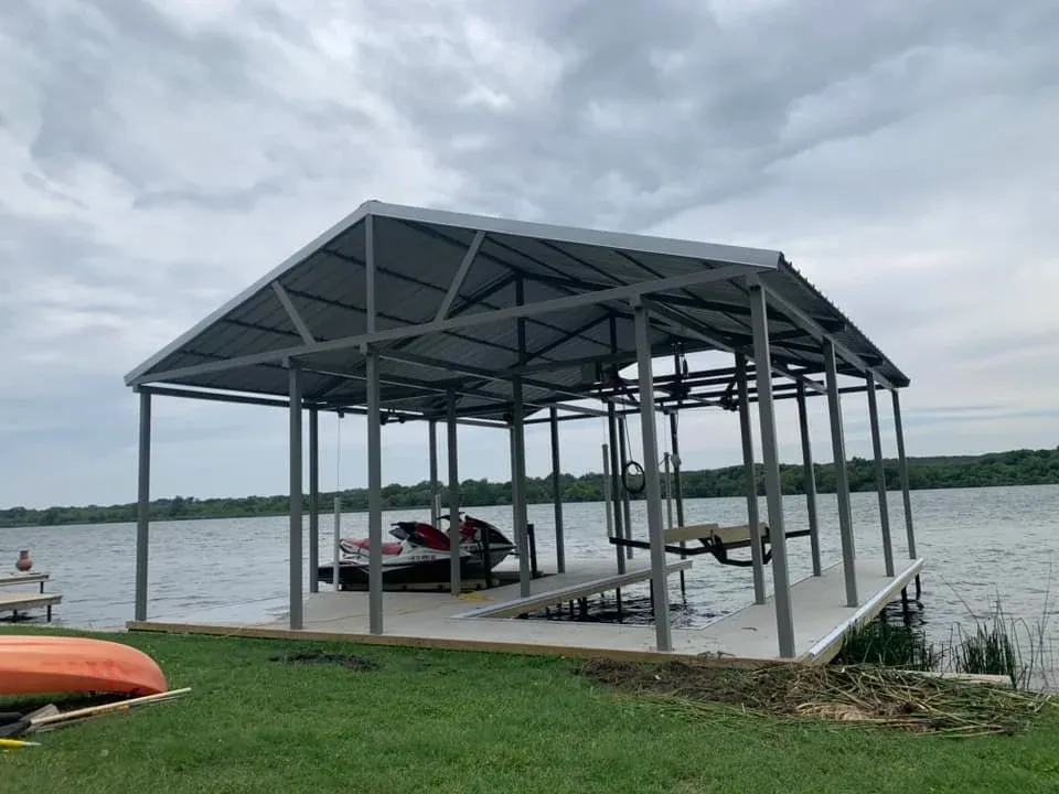 Open steel boat cover over a dock with jet skis on a grassy shoreline